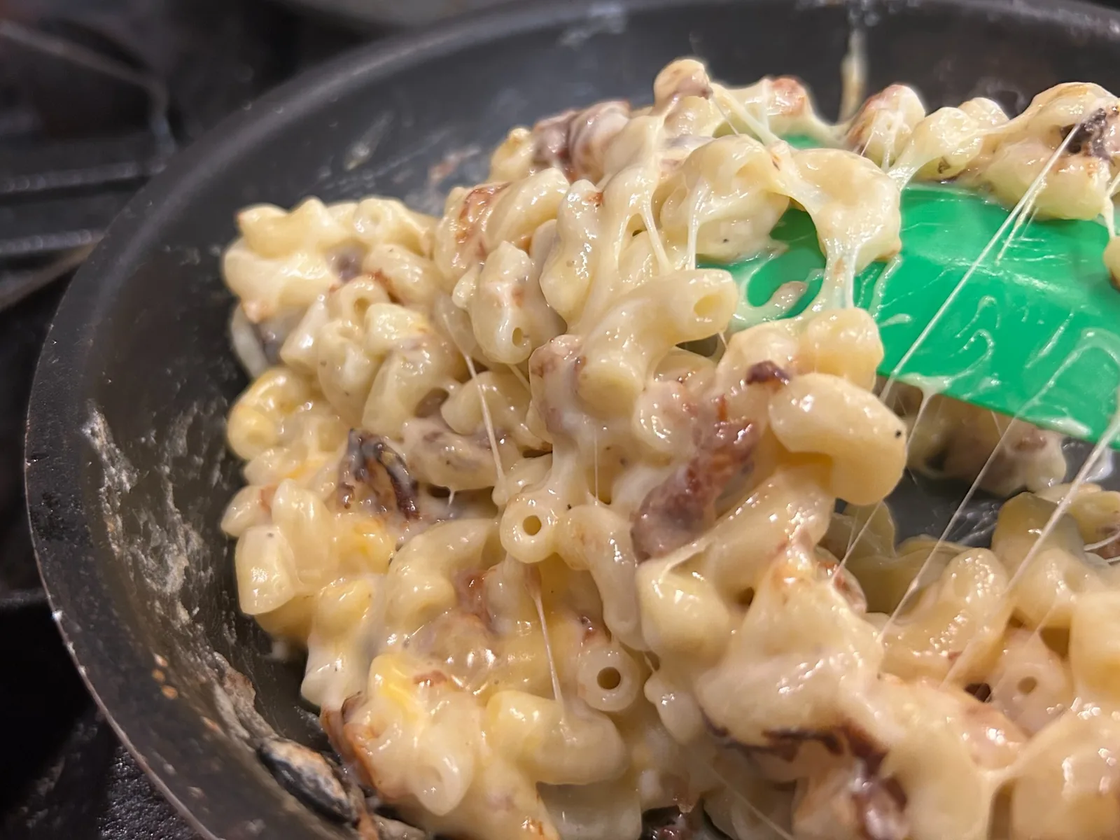 Homemade macaroni and cheese at Kat’s Café & Restaurant being made on the stove, lifted from a skillet with a green spatula, showing strands of melted cheese stretching between the pasta.