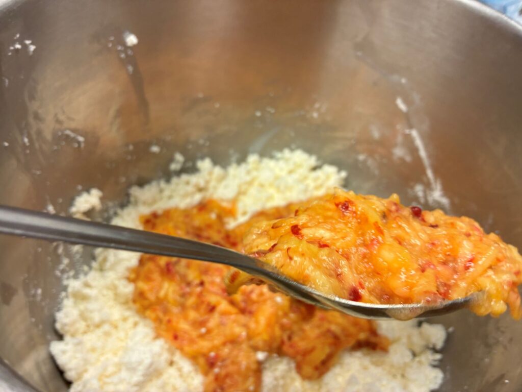 Shredded Peach Puree Filling on a Large Stainless-Steel Mixing Spoon Being Added to a Stainless-Steel Mixing Bowl Having Farmers Cheese Already There on the Bottom.