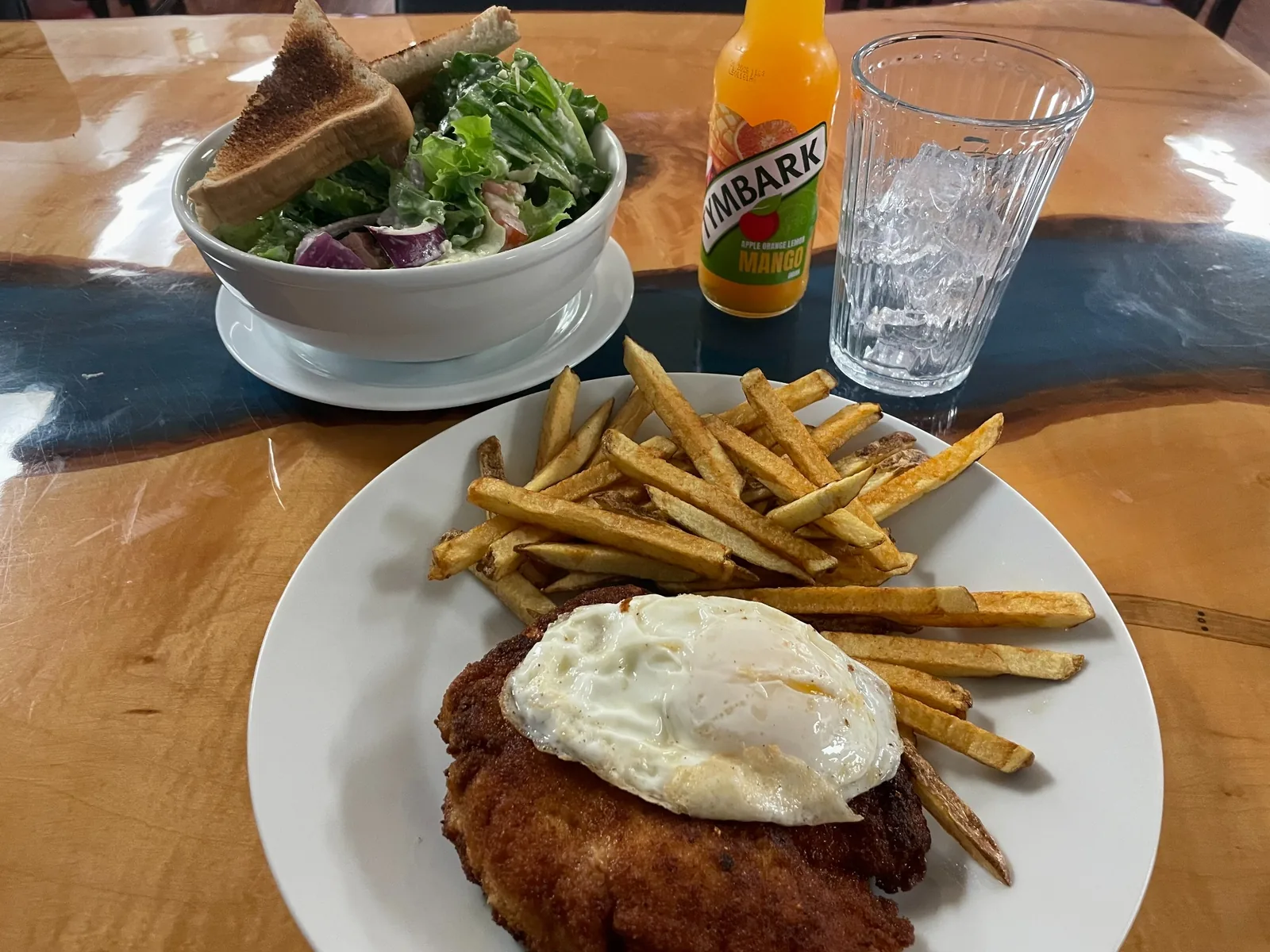 Chicken cutlet at Kat’s Café & Restaurant, topped with a fried egg, with hand-cut fries, a house salad with toast, and a bottled fruit drink on a wooden table.