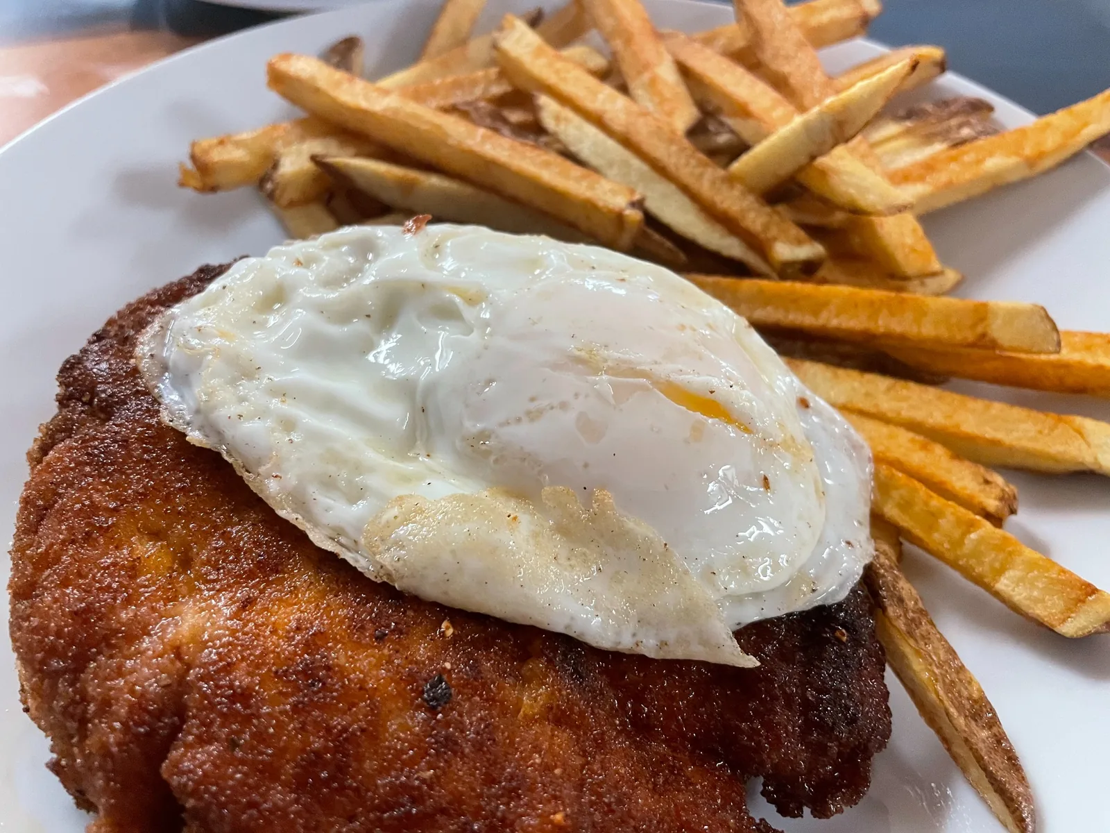 Close-up of a golden chicken cutlet at Kat’s Café & Restaurant, topped with a fried egg, served beside hand-cut fries on a white plate.