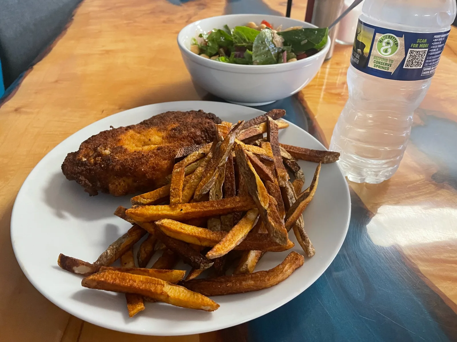Breaded chicken cutlet and hand-cut fries on a white plate, with a house salad and bottled water behind it on a wooden table at Kat’s Café & Restaurant.