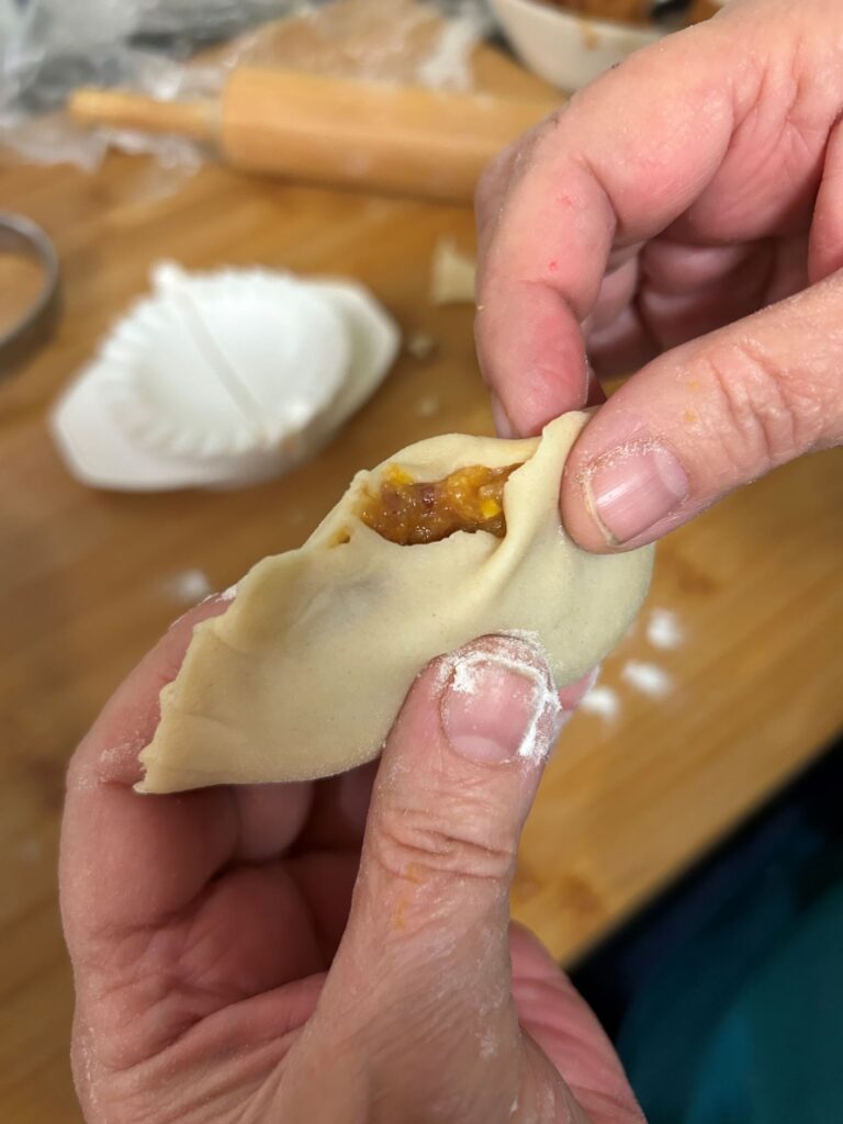 A photo of a Pilgrims Pierogi in the last stages of it being hand-crimped, showing the pierogi held in a female hand with fingers from the other hand finishing up the crimping process, with some of the filling visible before the pierogi is fully crimped, having a pierogi workstation in the background.