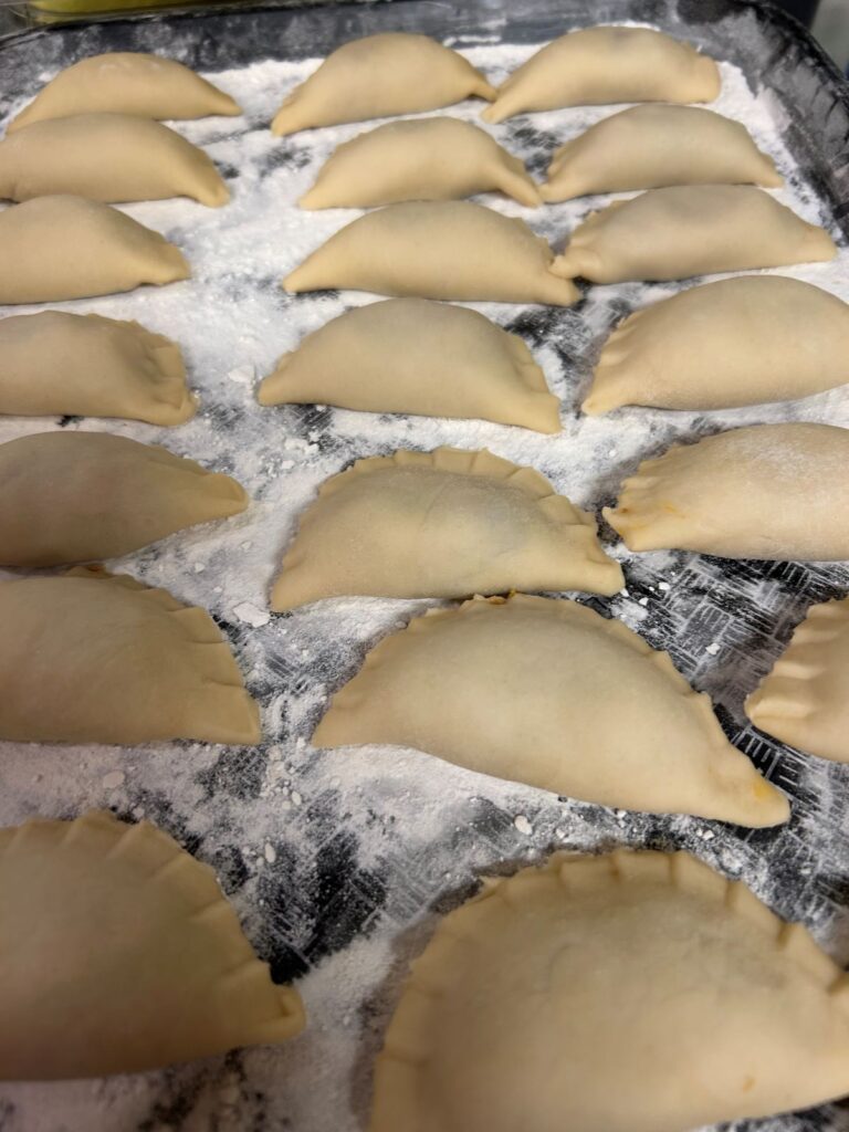 A photo of Kat's Pilgrims Pierogis Freshly Made and Laid Out on a Black Tray Sprinkled with White Flour.