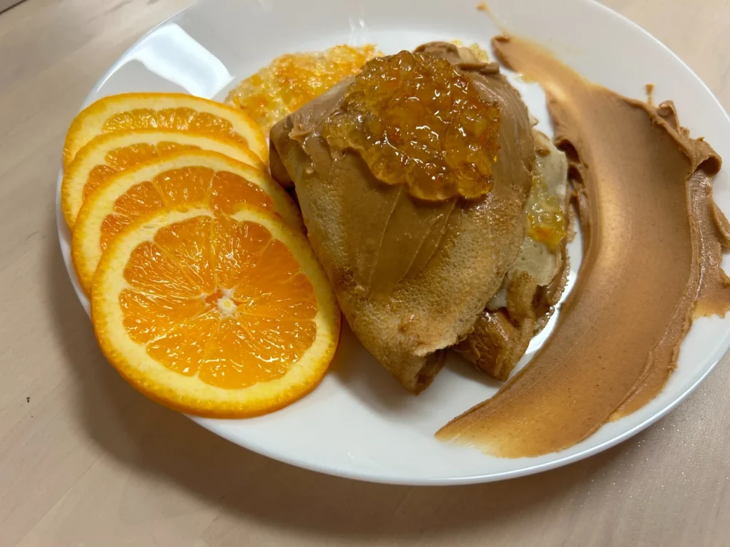 Folded PB&J crepe at Kat’s Café & Restaurant, topped with peanut butter and jam, served with orange slices and a sweep of peanut butter on a white plate.