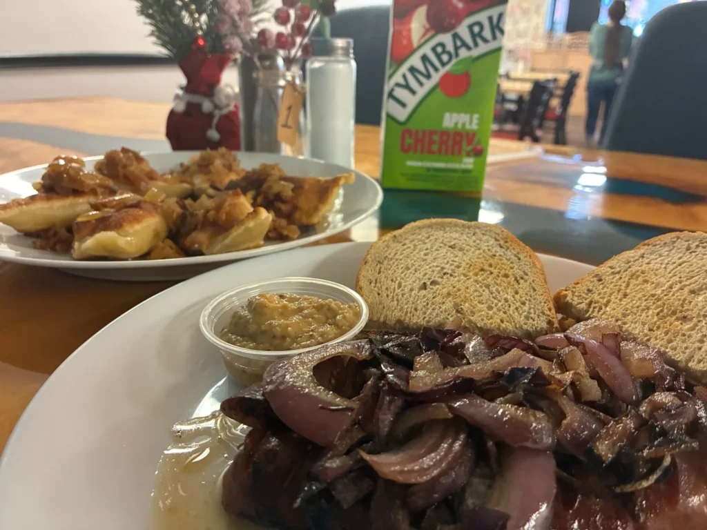 Pan-grilled Polish kielbasa topped with onions, served with bread and mustard, with pan-fried pierogies topped with caramelized onions on a separate plate behind it at Kat’s Café & Restaurant.