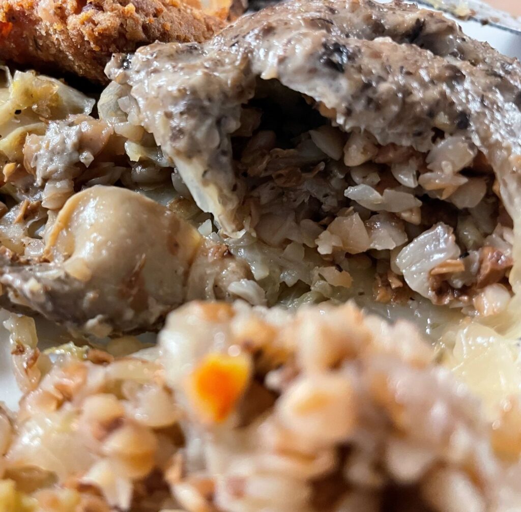 Kat's Buckwheat Mushroom Halupkies inside close up showing the vegetarian filling spilling out of the sauteed cabbage leaf wrap and topped with homemade wild mushroom sauce, served on a white plate next to fried fish and łazanki.