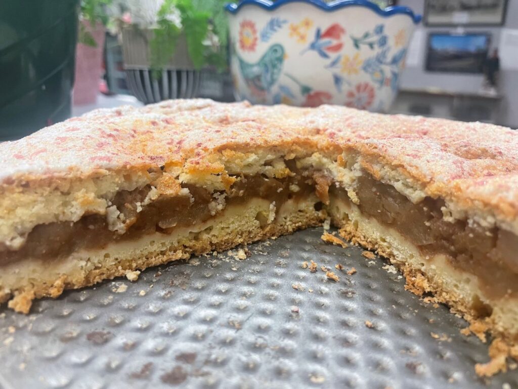 Homemade Apple Pie Freshly Cut showing the natural filling and buttery crust, lightly coated with real powdered sugar and pinkish sugar crystals on a grooved metal tray, seeing the cafe background including fresh flowers and plants and an artisan bowl.