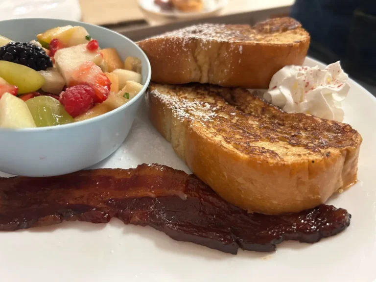 Thick slices of French toast at Kat’s Café & Restaurant served with maple-glazed bacon, whipped cream, and a bowl of fresh fruit salad.