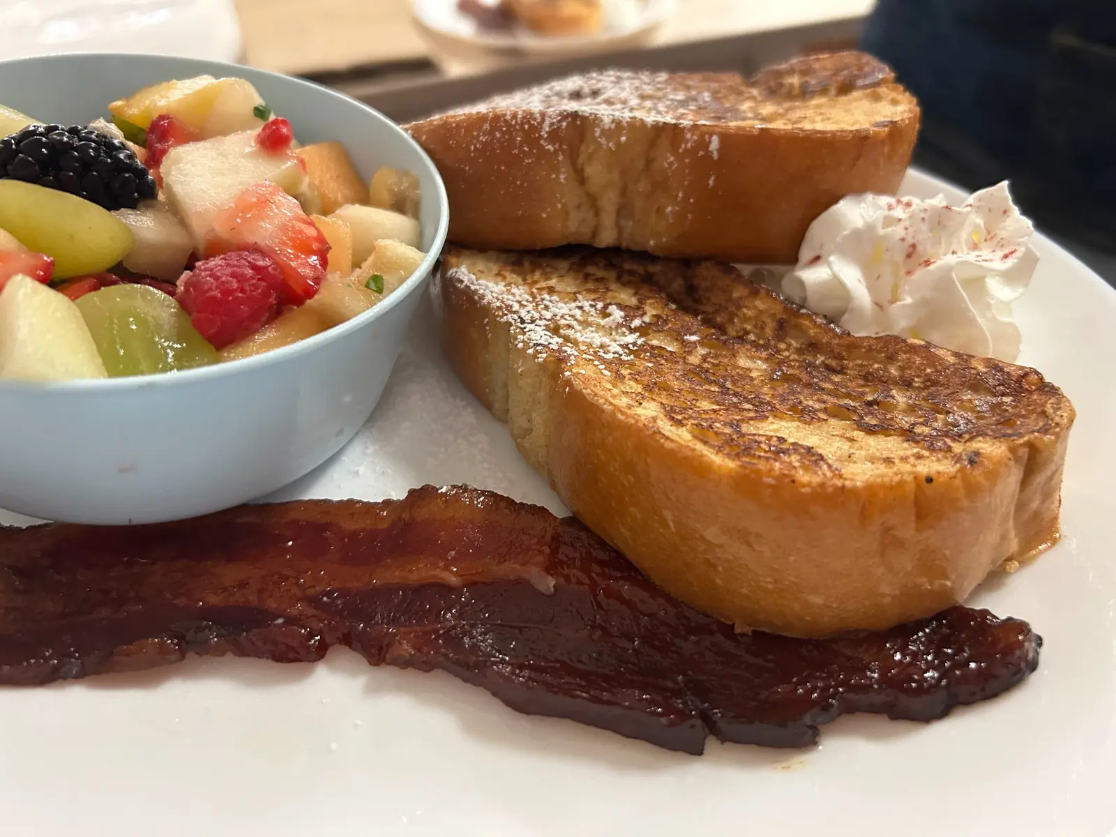 Thick slices of French toast at Kat’s Café & Restaurant served with maple-glazed bacon, whipped cream, and a bowl of fresh fruit salad.