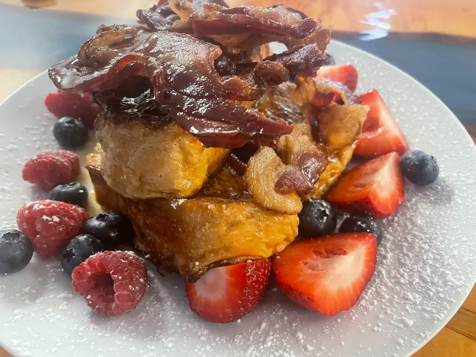 Stacked French toast topped with maple-glazed bacon and surrounded by strawberries, blueberries, and raspberries on a white plate at Kat’s Café & Restaurant.