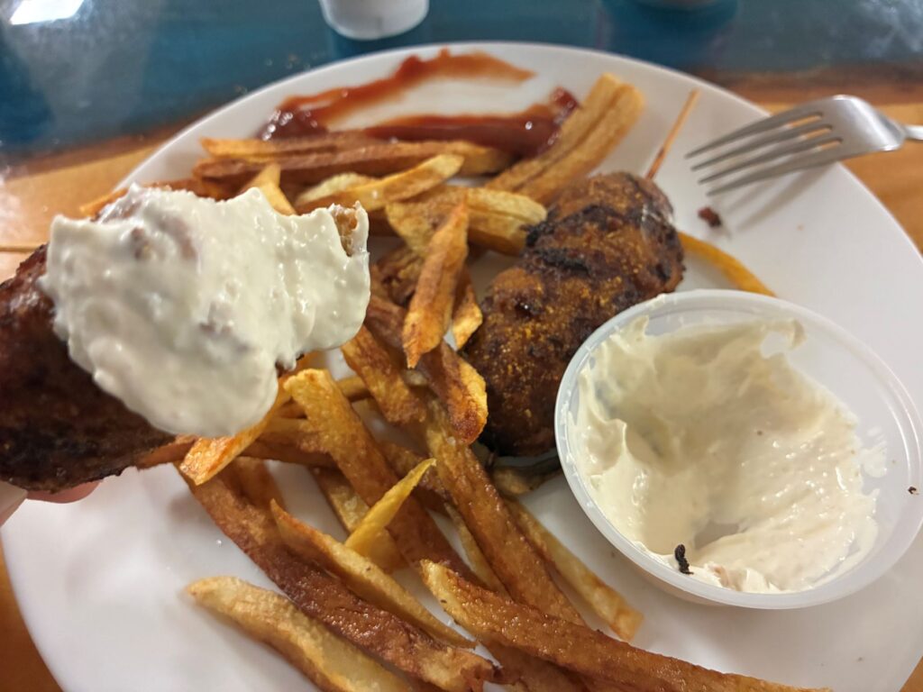 A Bite of Kat's City Chicken with Homemade Dip on It with the Rest of the Appetizer on a white plate below having Fresh Cut Fries, Another Piece of City Chicken on a Stick, Smeared Ketchup, with a fork propped up against the plate, served on the maple tiger wood dining table at the restaurant.