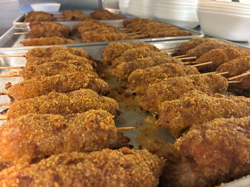 Trays of City Chicken in a Commercial Kitchen Setting Having White Plates and Bowls Stacked to the Side of the Metal Baking Trays Completely Full of Rows of City Chicken.
