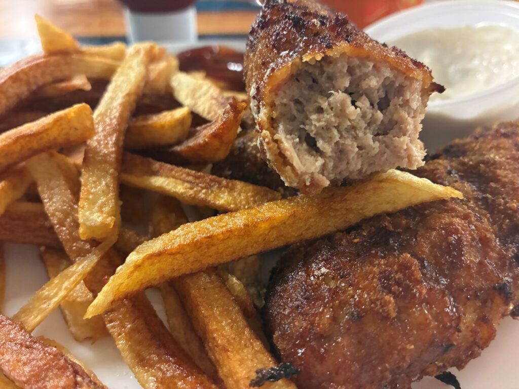 A piece of Kat's City Chicken Partially Eaten Showing the Moist Ground Meat Inside and Crispy Lightly Breaded Outside on Top of the Appetizer Meal having Fresh Cut Fries with Ketchup and Garlic Parmesan Dip in a Container, all served on a white plate on the custom river run live edge wood table at the restaurant.