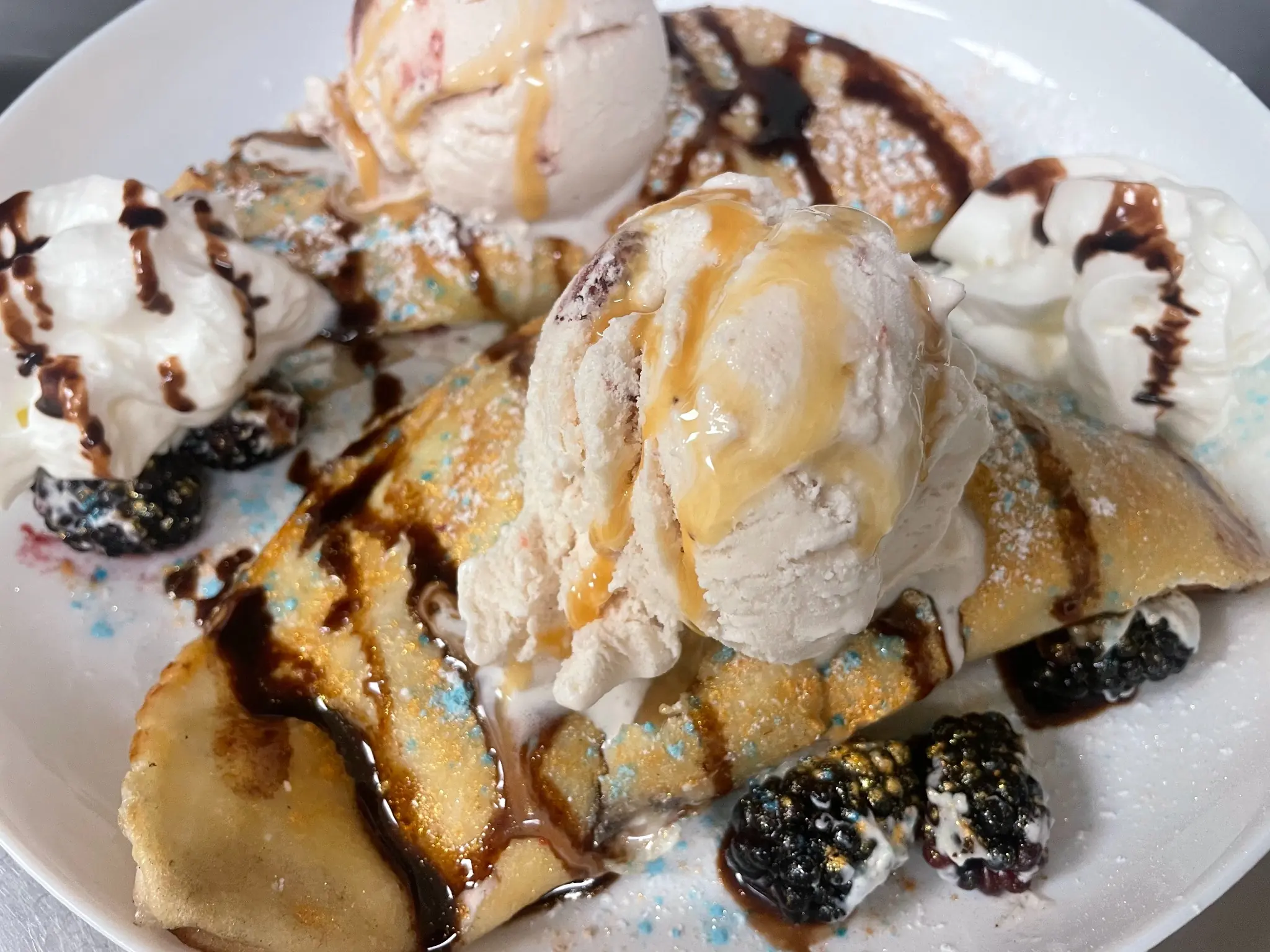 Polish-style sweet cheese crepes topped with ice cream and caramel sauce, served with blackberries, whipped cream, powdered sugar, and chocolate drizzle.
