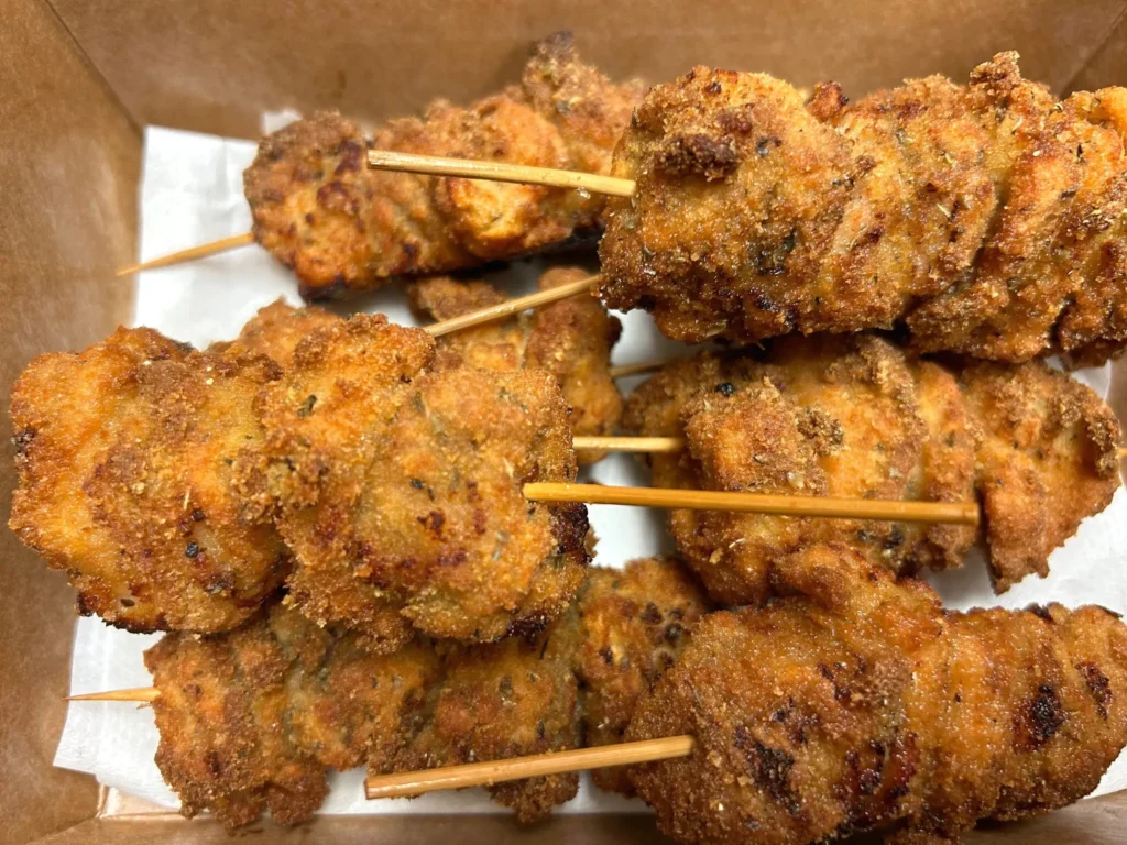 Close-up of crisp, golden-brown chunk-style City Chicken pieces arranged on wooden skewers inside a Kat’s Café takeout box.