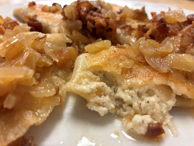 Kat's Classic Potato Cheese Pierogi Topped with Sauteed Onions and Real Crumbled Bacon Having a Half Cut Pierogi in the Front and Center Showing the Fresh Filling on the Inside, served on a white plate.