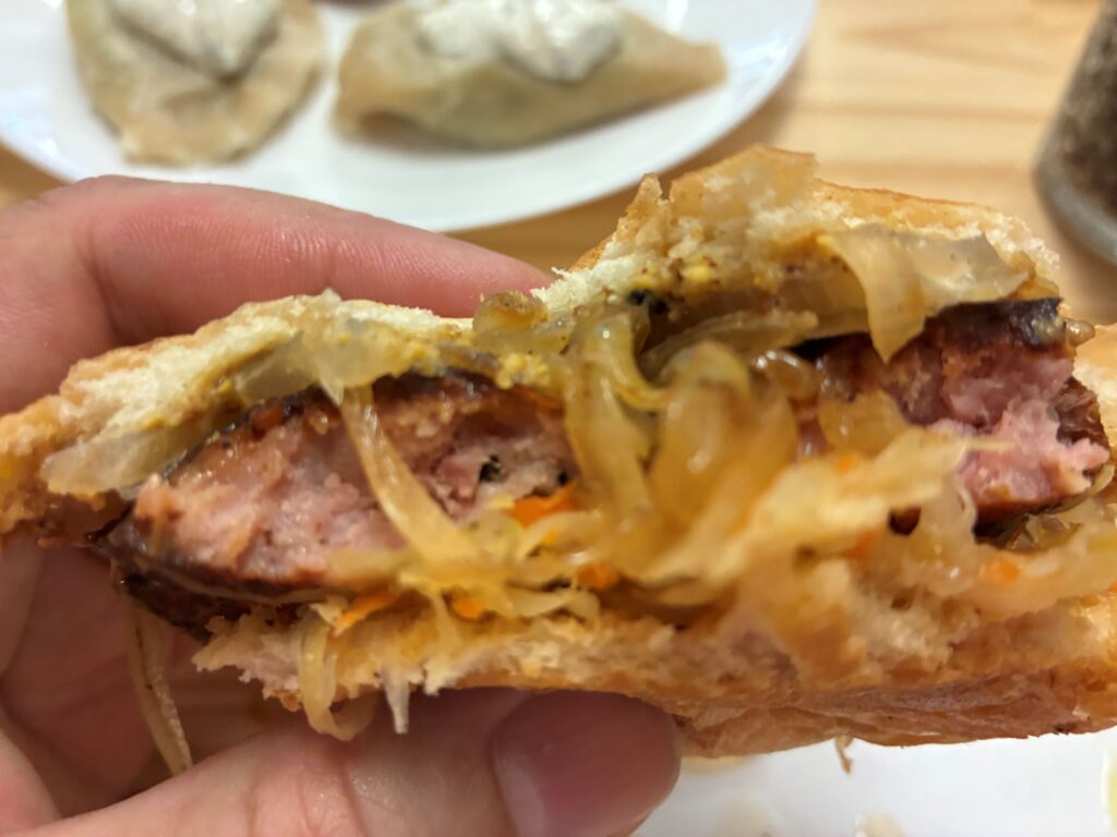 Kat's Kielbasa Sauerkraut Hoagie Partially Eaten Held by a Male Hand with Pierogies on a White Plate on a Light Wood Table in the Background.
