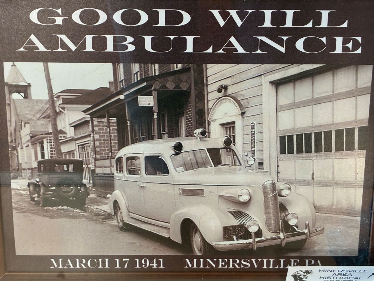Historical Photo from March 17, 1941, of a Goodwill Ambulance parked in front of a fire station garage door with a soft top vintage automobile parked behind it in the opposite direction in front of a brick house with a porch.