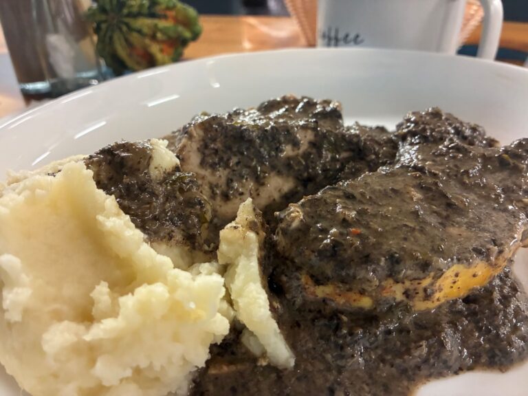 Kat's Creamy Chicken Wild Mushroom Dinner served on a white plate having pieces of tender chicken breast covered in a wild mushroom sauce with a side of mashed potatoes, in the background a white mug and a green-orange pumpkin squash on the custom ambrosia table at the restaurant.