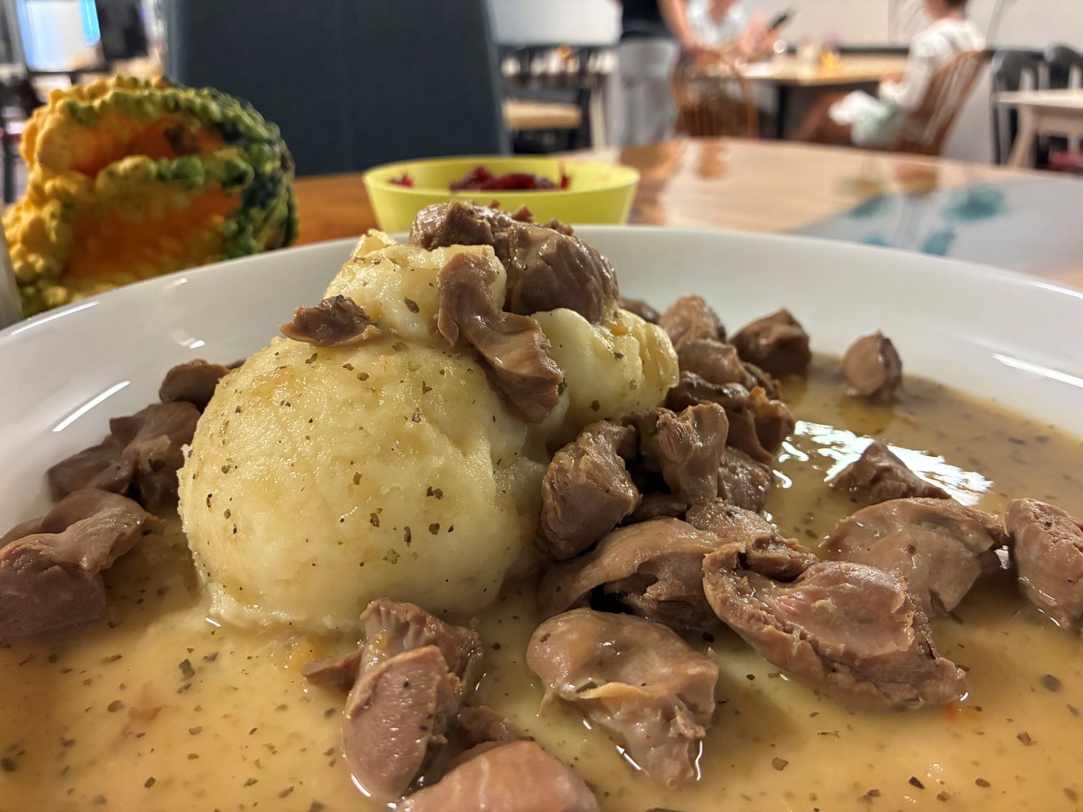Chicken Gizzards in Sauce and Mashed Potatoes served on a large white plate with an upward beveled lip, having a yellow bowl with shredded beets and apples on the side, on an ambrosia maple wood table surface, with a young couple being served sitting on one of many ash wood tables in the background.