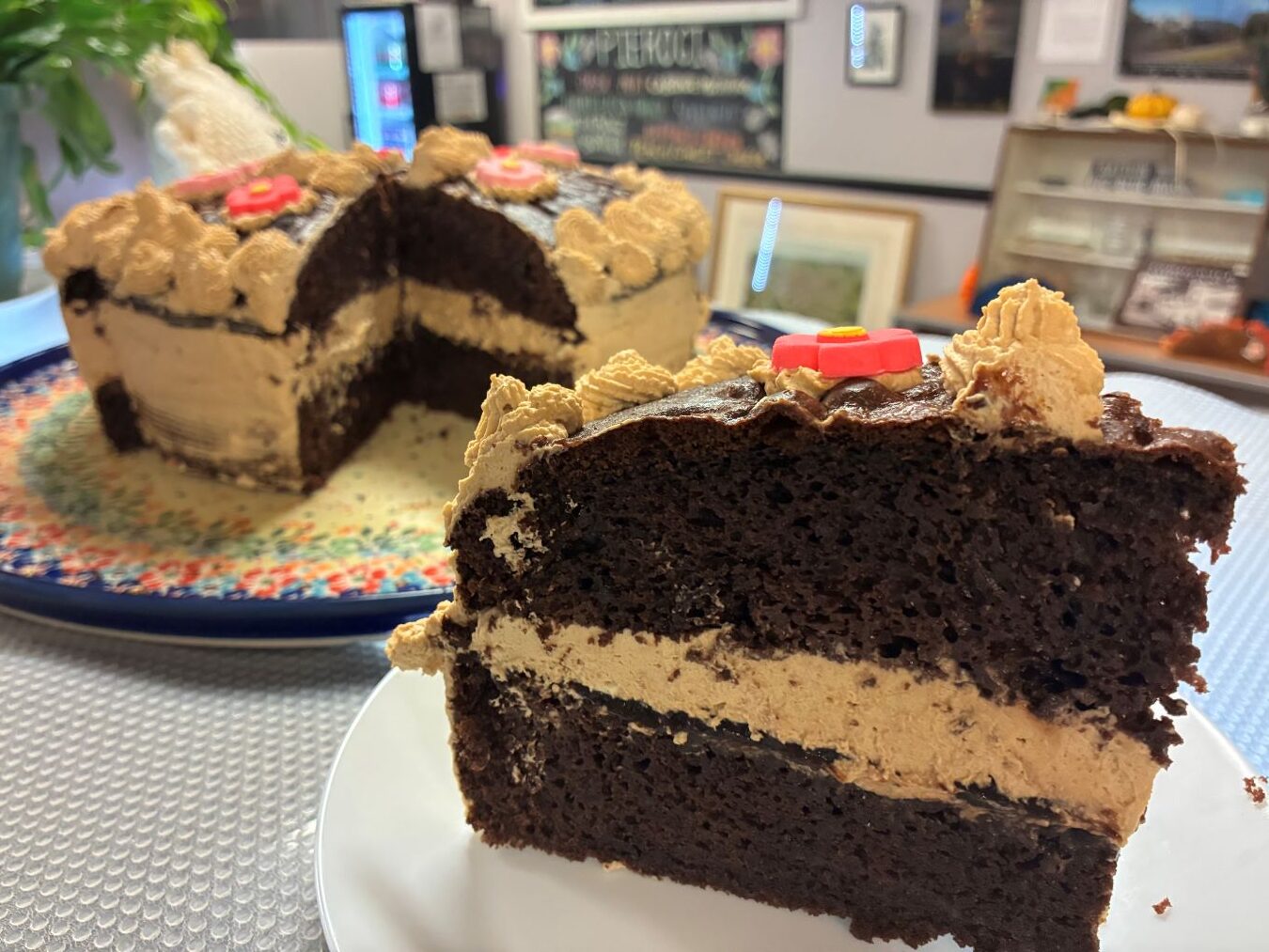 Kat's Chocolate Coffee Creme Cake Slice in the Front Center with the Rest of the Cake Along with the Background of the Restaurant in the Front Area in the Background.
