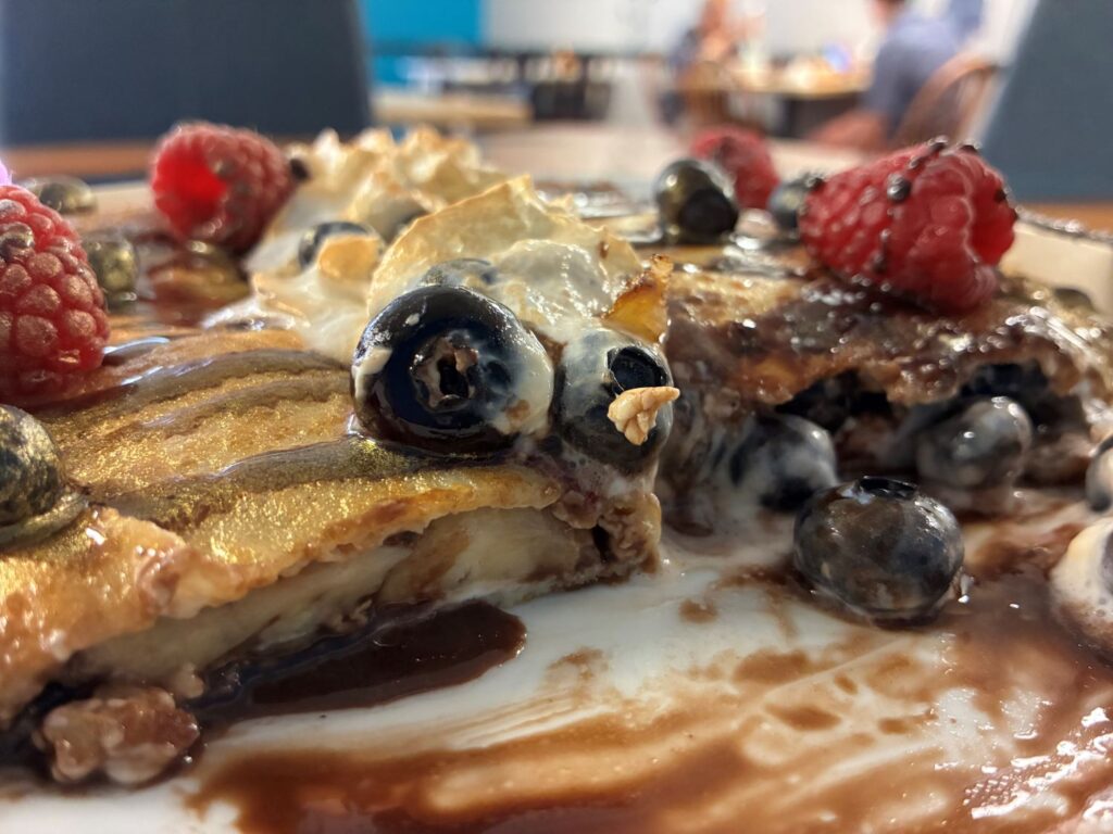 Two blintzes, also known as Slavic or Polish crepes, served on a white plate having chocolate, Nutella, and sweet cheese banana and berries filling coming out of the ultra-thin folded blintz pancake-like-wrap dusted with gold and colored flavoring, covered in raspberries and blueberries with puffs of whipped cream, seeing an older lady with a younger gentleman having a meal seated at a table in the restaurant in the background.