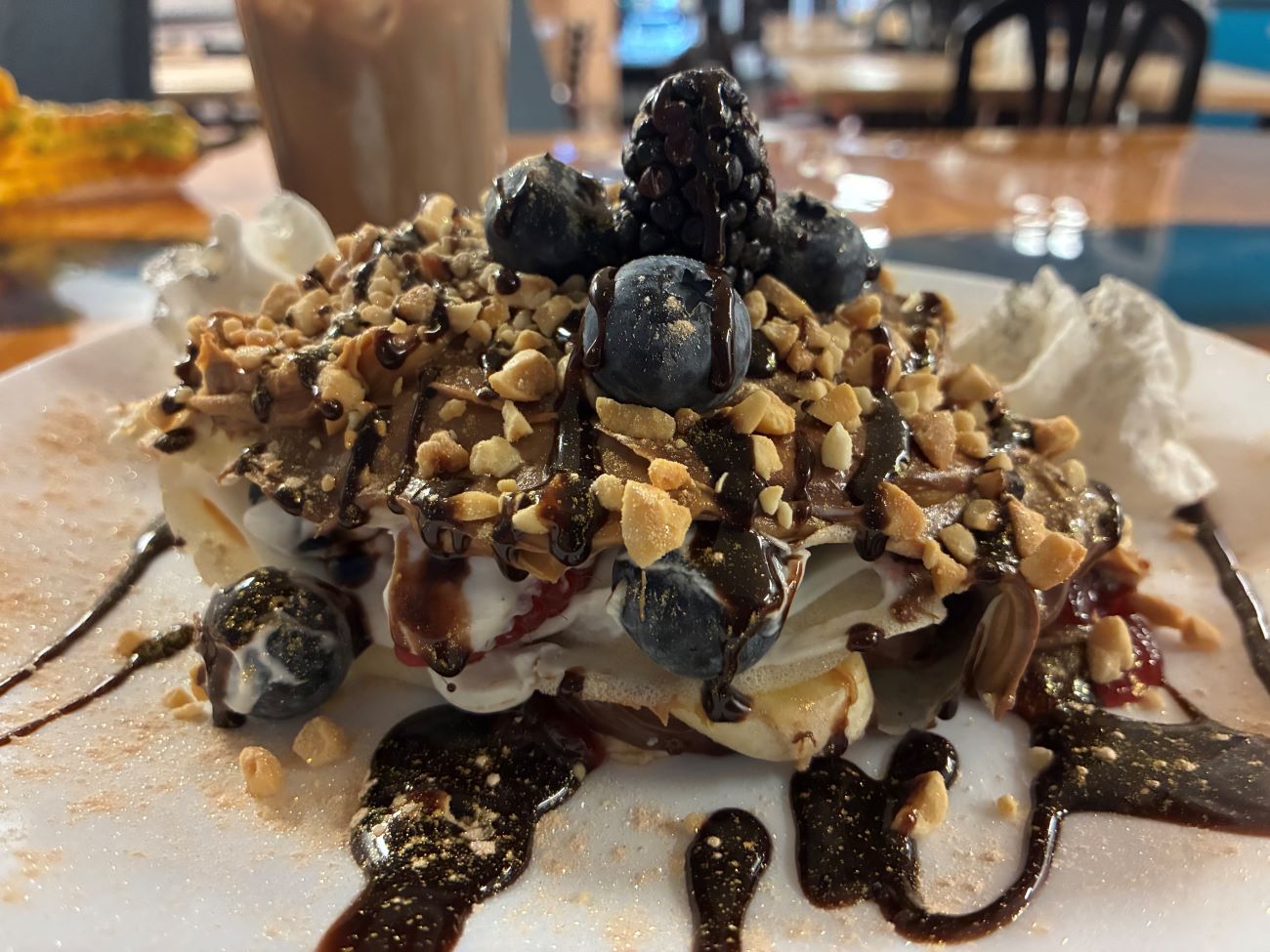 Everything Crepe-Blintz Loaded with Peanut Butter, Nutella, Sweet Cream, Chopped Nuts, Blueberries, Blackberries, Natural Jam, Whipped Cream, and Chocolate Sauce, served on a white plate with Iced Coffee.