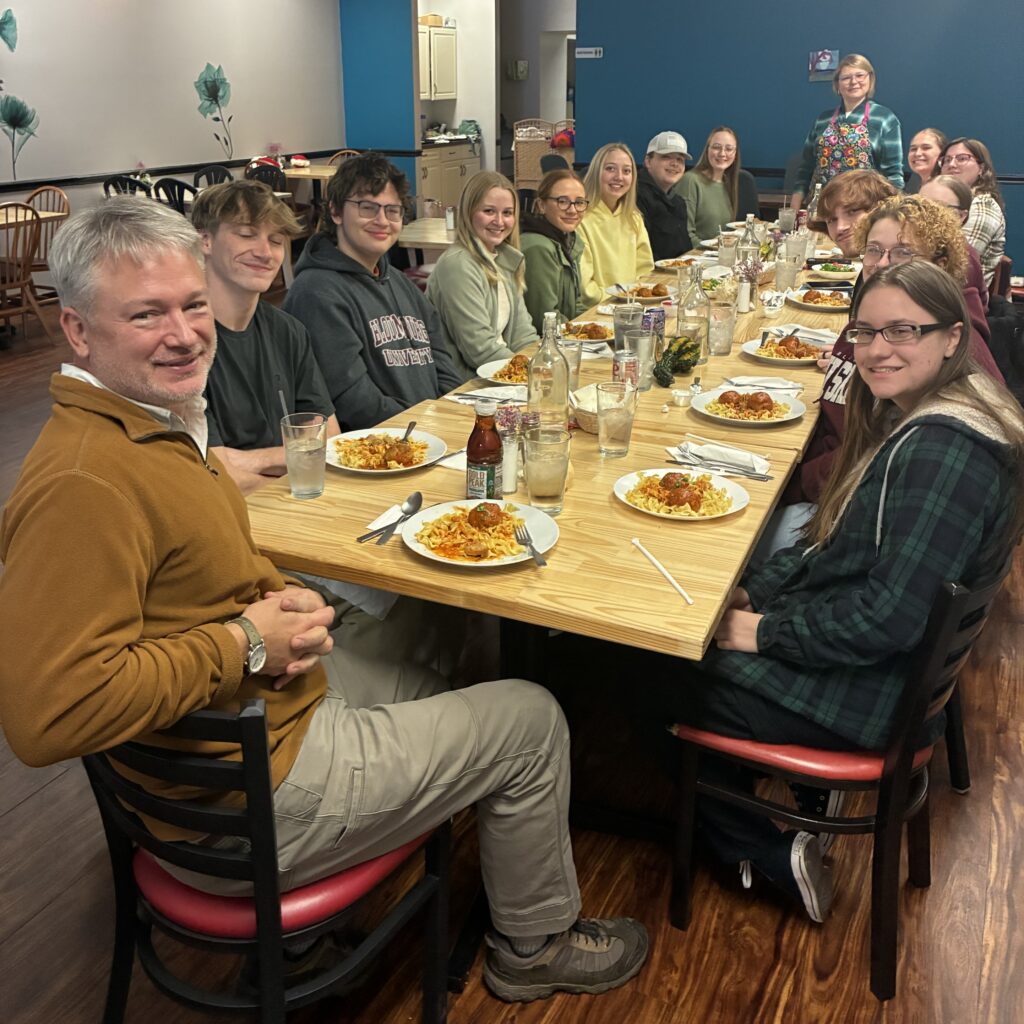 A photo of young adults from a Pennsylvania college with their professor seated around a table for a lunch event hosted at Kat's with the chef and owner standing at the far end of the table smiling.