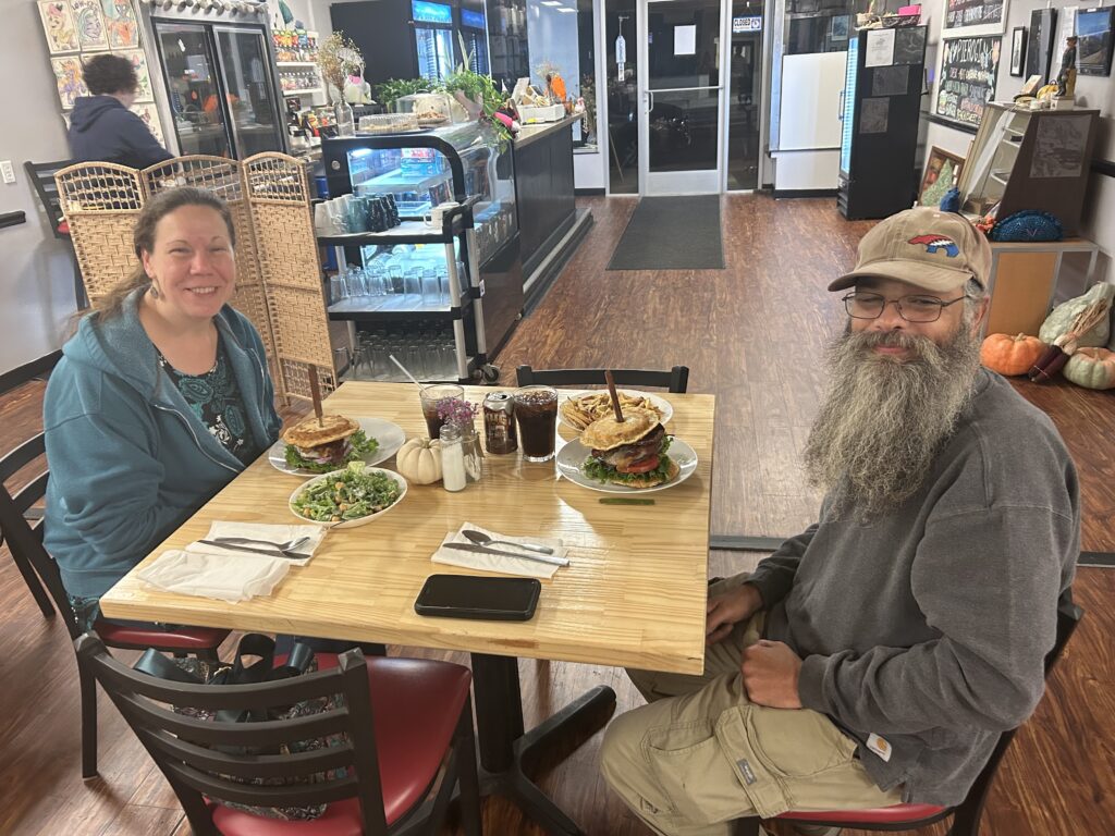 A happy smiling couple seated across from one another both having pierogi burgers on their plates looking up with the front area of the cafe-restaurant in the background.