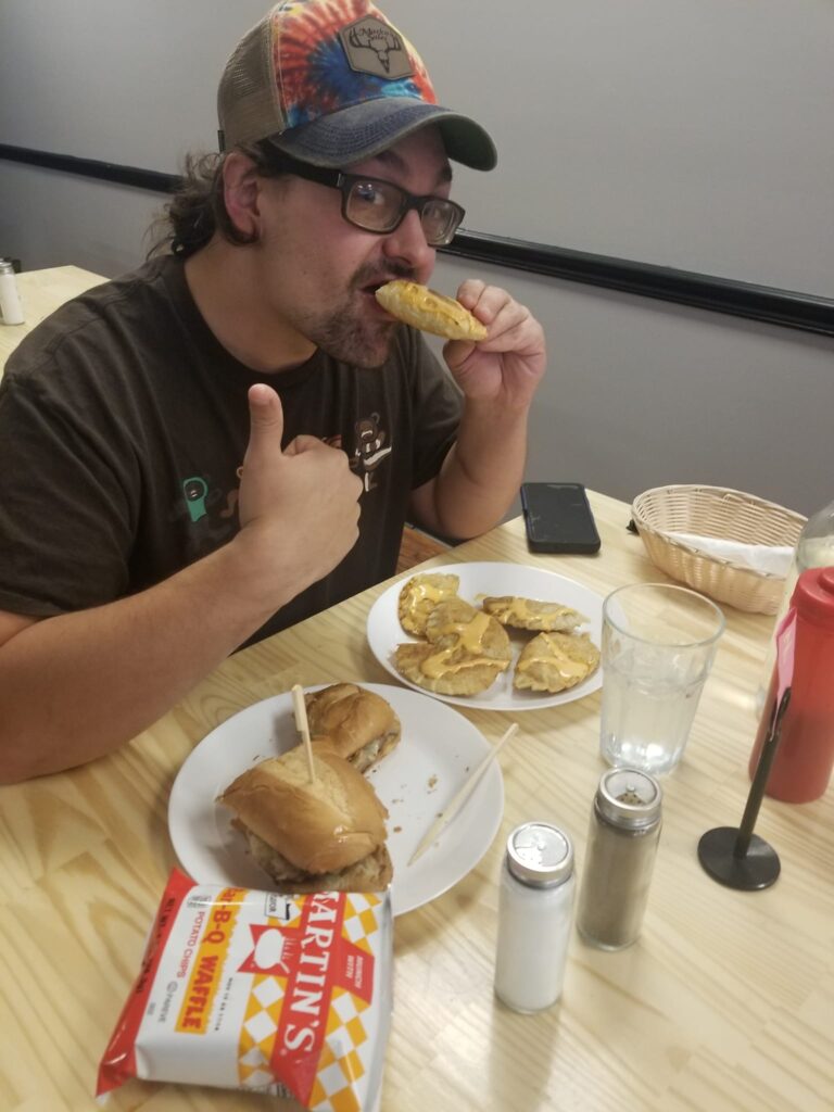 A photo of male customer holding up a pierogi to his mouth in the process of eating it while giving a thumbs up with his other hand and having a plate of pierogies and a half-finished sub on a plate on the table with a bag of chips and other items like ketchup, salt and pepper shakers, and clear glass of water.