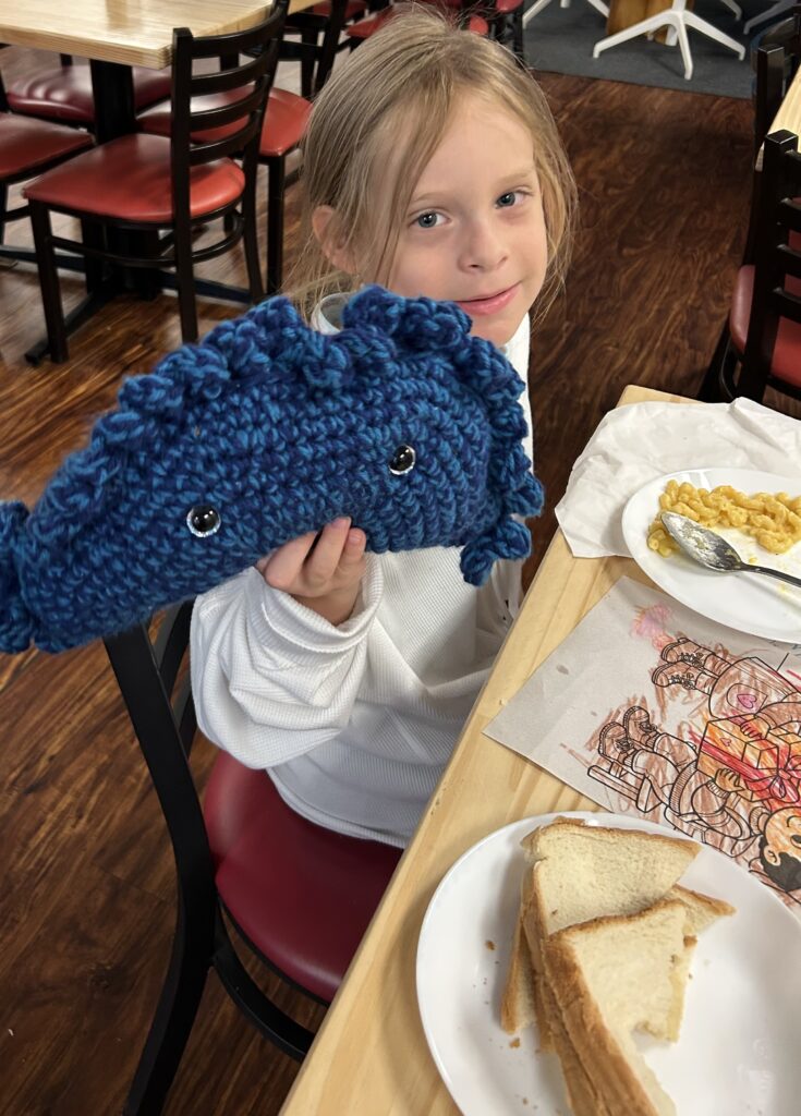 A photo of a young girl holding up a blue pierogi pillow with one hand while seated at a table having a page from a coloring book and food on plates in front of her.