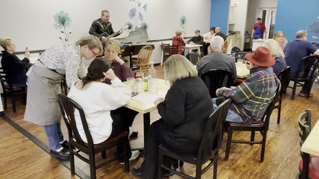 A photo of lots of different customers sitting at tables eating their meals with a local musician playing acoustic guitar in the background.