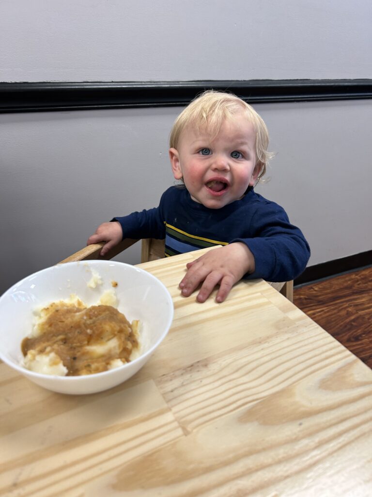 A photo of a small boy seated at a table with a bowl of food in front of him looking up and visibly happy.