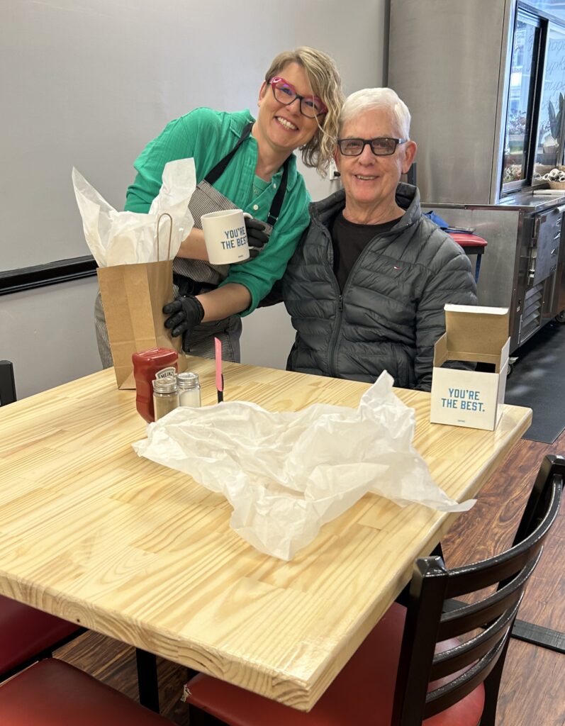 A photo of Kat standing next to seated white-haired gentleman and leaning in towards him while holding a white coffee mug with light blue letters "You're the Best" and a gift bag with white wrapping paper sticking out of it with the same white wrapping paper out on the table with an open box having the same words as on the cup.