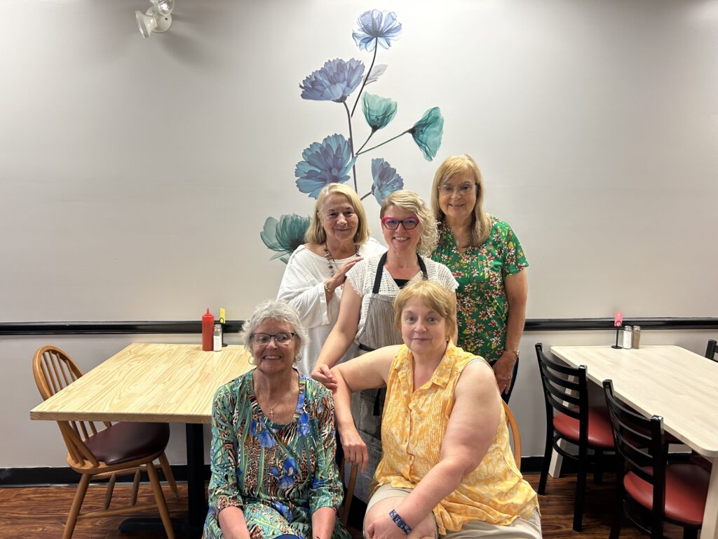 A photo of a group of ladies some seated while others standing with Kat, the owner of the restaurant, in the middle of the group and in the background clean, empty ash wood tables with red and black chairs and a wall with a visible flower painted on the wall.