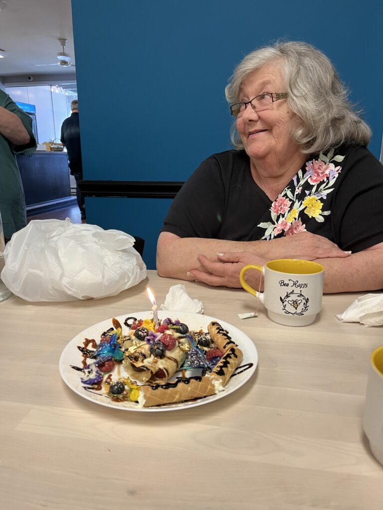 A photo of a smiling female customer in eyeglasses looking up at someone not in the photo who just served her an everything blintz for her birthday with a white and yellow cup also in front of her on the table with a bumble bee and the words "Be Happy".