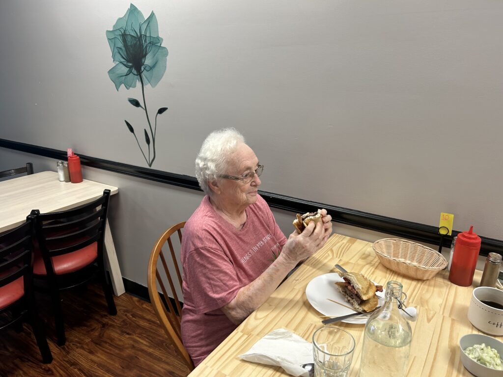 A photo of an older lady with white hair holding a pierogi burger in both hands while looking down at it about to take another big bite of it with the other half on the white plate in front of her on the ash wooden table that she is sitting at in the restaurant.