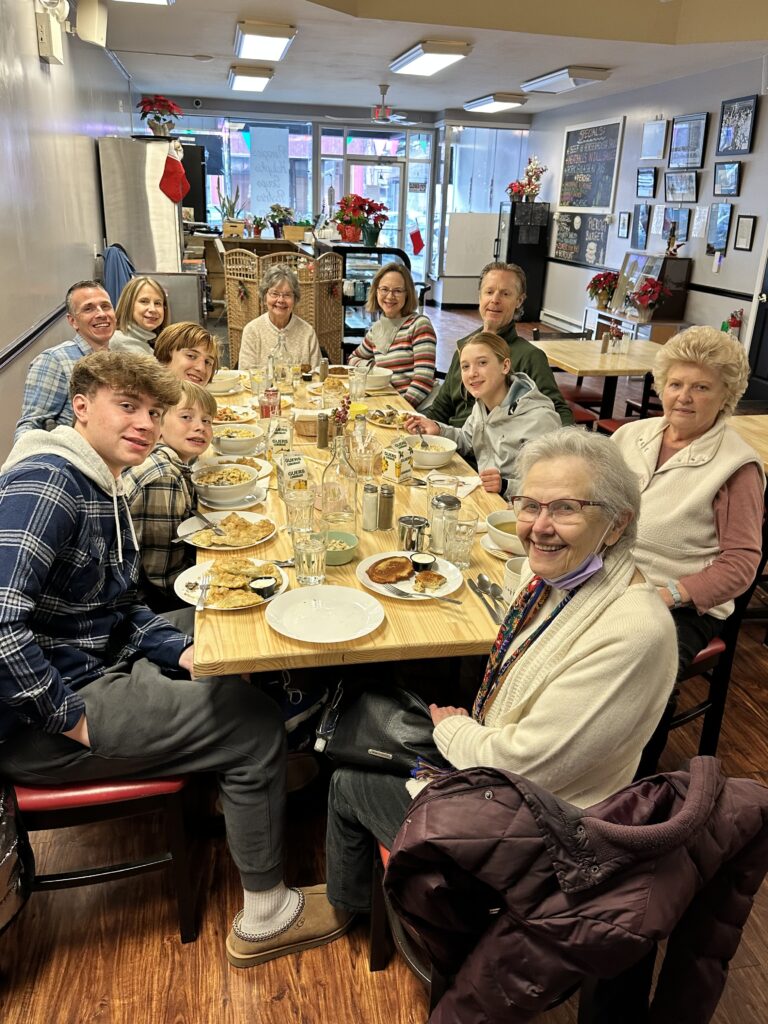 A photo of a multi-generational group of people from young to old all sitting around a bunch of joined tables filled with plates, glasses, utensils, and food, with the front of the cafe-restaurant in the background also seeing out into the main street out front of the establishment through the floor-to-ceiling glass windows.