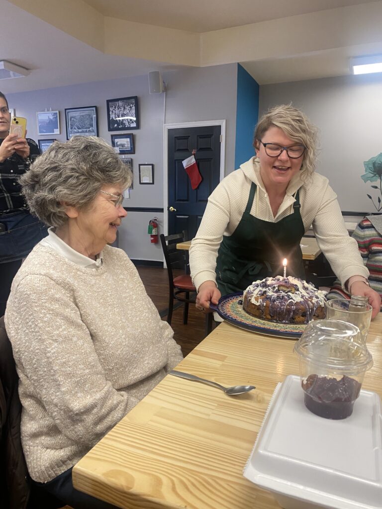 A photo of a Kat placing a birthday cake with a single lit candle in the center of the cake down on a table at the restaurant in front of a smiling elderly lady looking down at the cake with the restaurant wall having pictures on it in the backdrop.