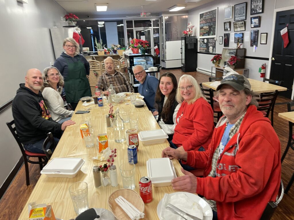 A photo of a group of customers casually dressed looking up and smiling sitting around an arranged long table with Kat standing the far-right corner wearing a chef's apron with the front area of the cafe-restaurant in the backdrop.