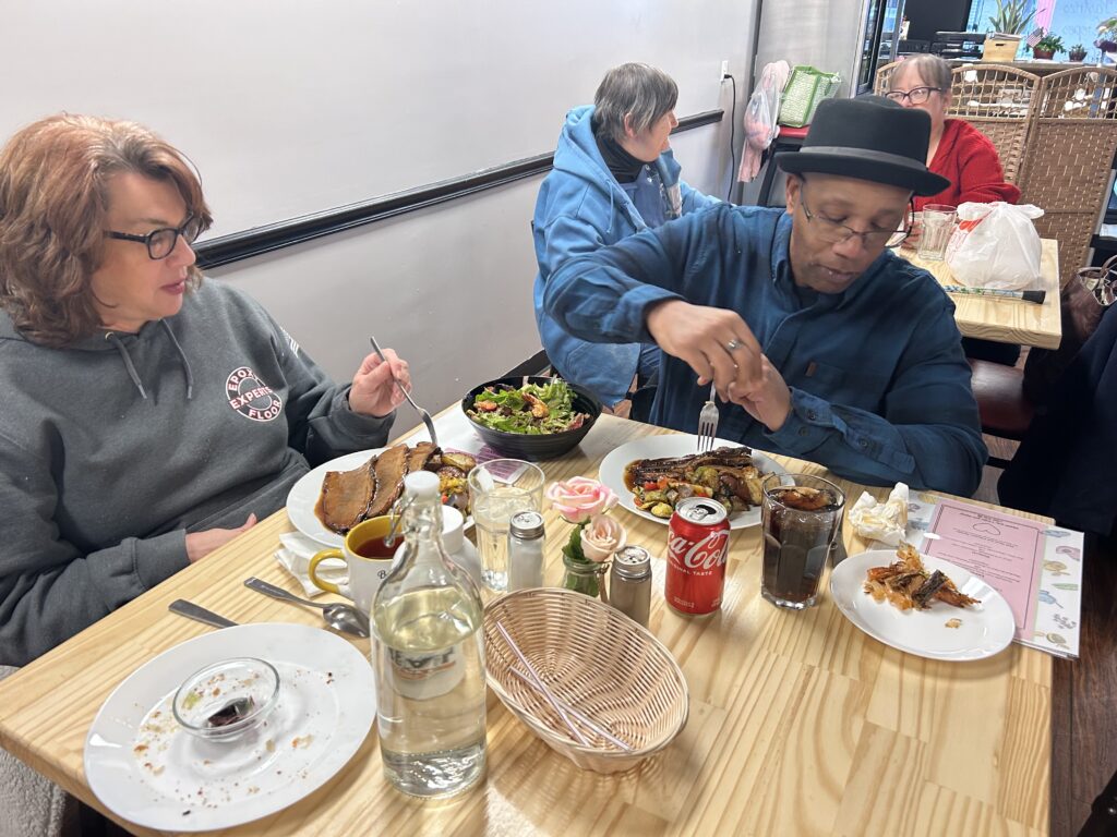 A photo of a couple seated at a table with the black gentleman looking down at his plate in the process of dipping his food into the sauce and the white lady holding a fork in the process of also eating both seated at a table filled with food and in the background another table with two females seated at it also having food on their table.