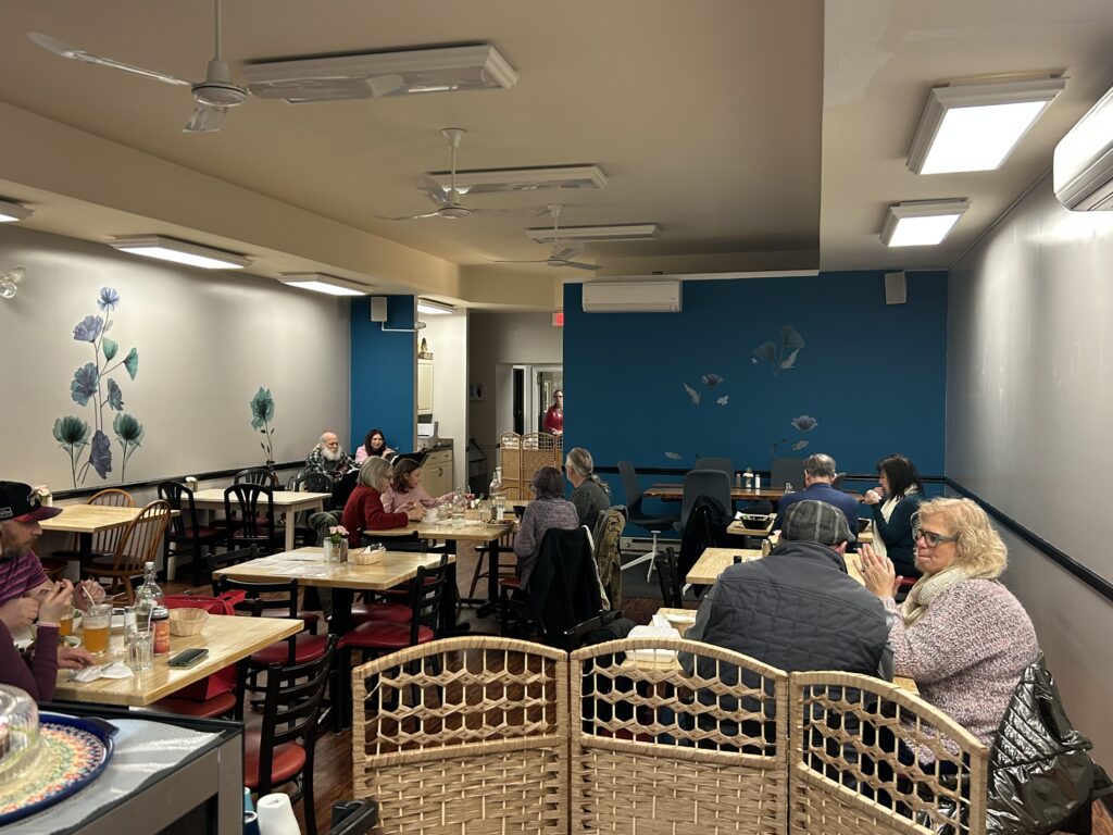 A photo of the dining area at the cafe-restaurant with customer seated at tables eating and in the far-left corner two local musicians seated while singing and playing instruments.