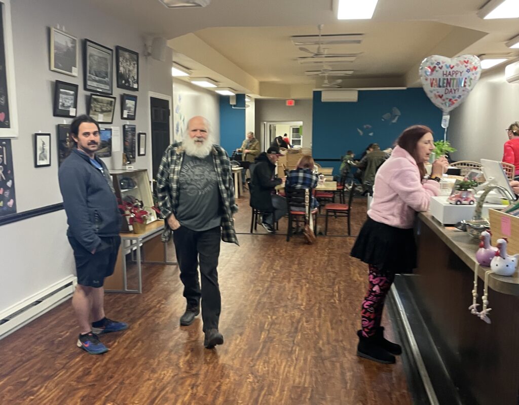 A photo of a customer walking directly ahead, another customer standing with his hands in his pockets also looking in the same direction, another customer leaning in towards the front counter and a dining area full of customers eating at Kat's.