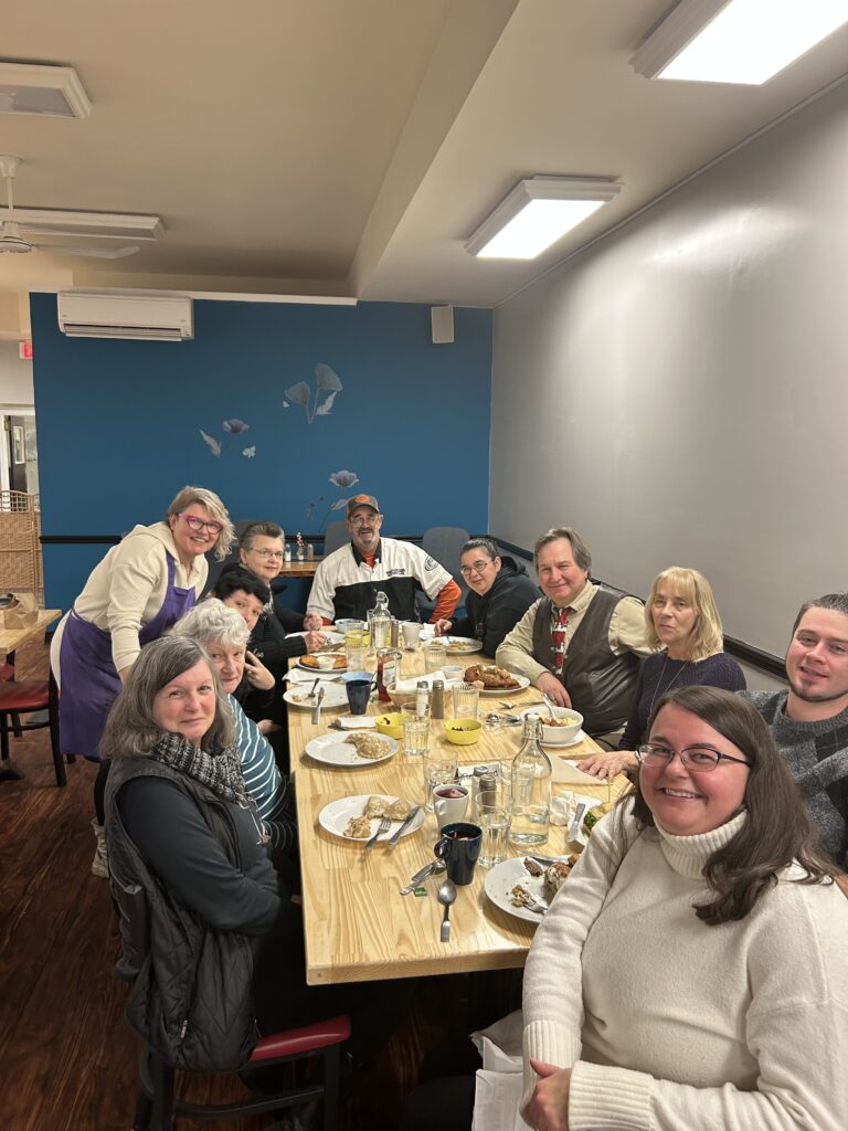 A photo of a group of happy customers of all ages sitting around a table with plates of leftover food from a huge meal and Kat leaning into the photo with everyone smiling.