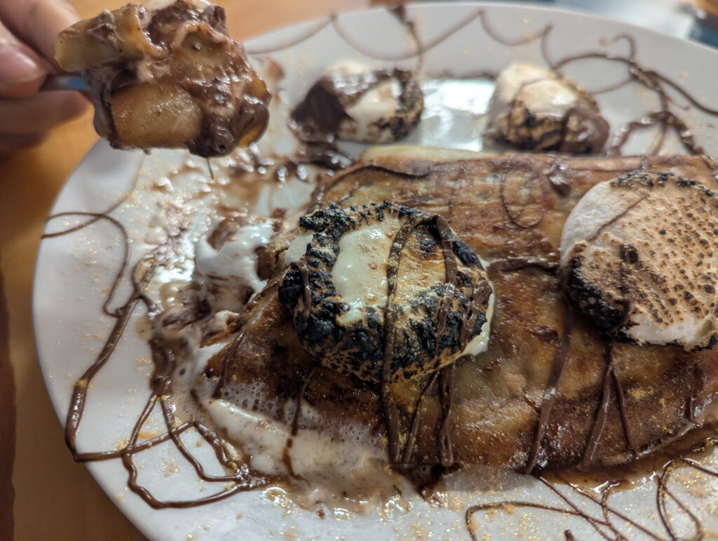 A photo of a Delicious Nutella S'mores Crepe with a piece of it on a fork showing the layered sweet goodness, served on a white plate garnished with chocolate swirls.
