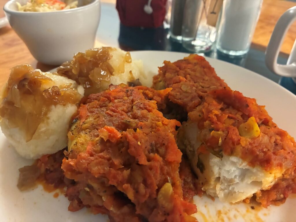 A photo of tender pan-seared white fish fillets topped with a hearty vegetable medley topped in homemade sauce slow-cooked from finely shredded carrot, celery root, and parsnip-combined with tomato paste to create a rich, savory layer that blankets the fish alongside two scoops of freshly made mashed potatoes covered in sautéed onions on a huge round white plate with a small bowl of spring Cole slaw.