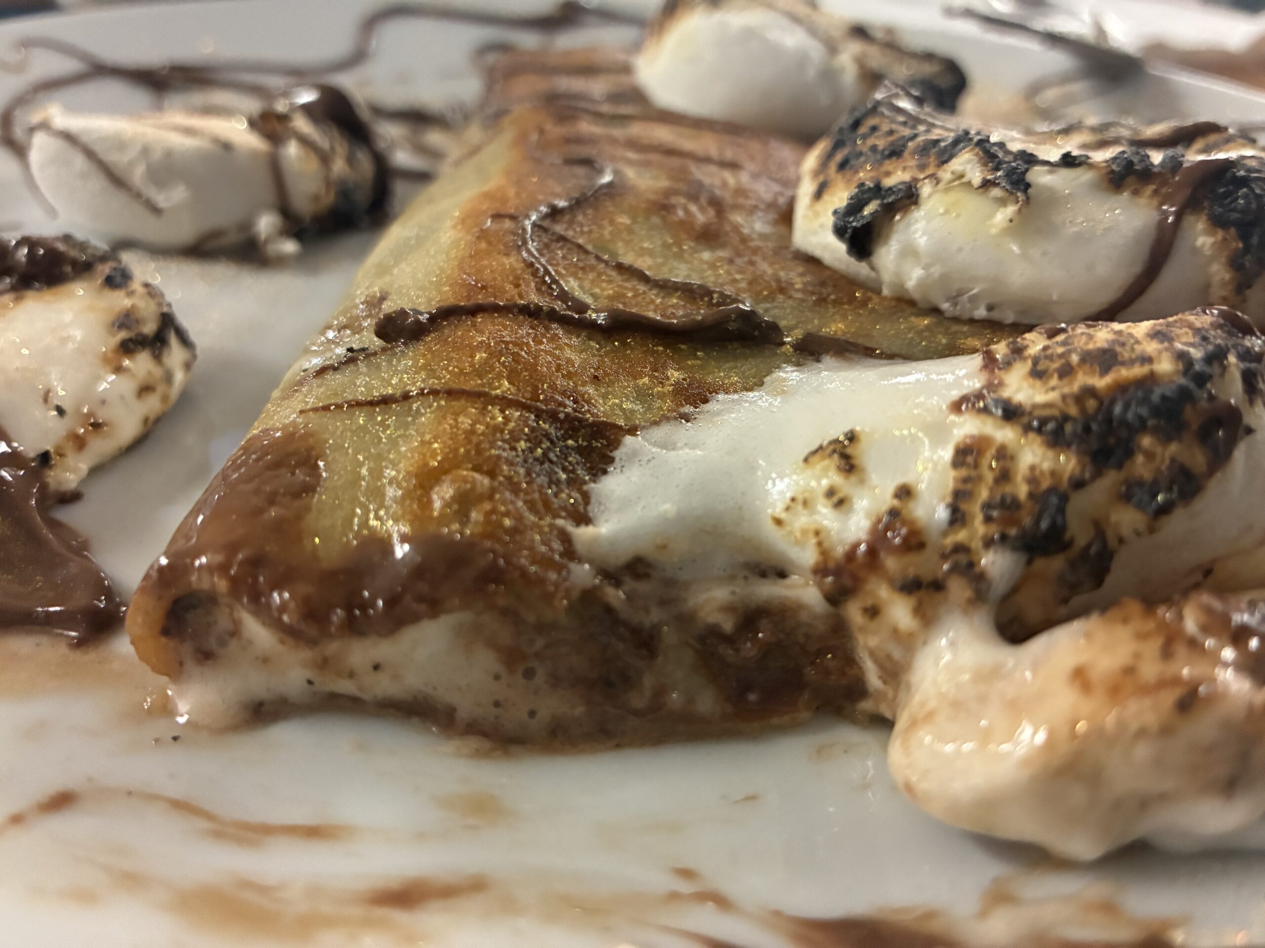 A photo of Kat's Nutella S'mores Crepe having a corner missing showing the inner layered hazelnut and crepe, with several toasted marshmallows on top of the crepe with real chocolate sauce swirls, served on a white plate.
