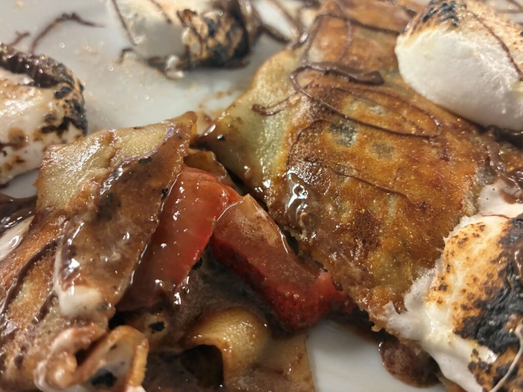 A photo of a Strawberry Nutella Smores Crepe cut in half with the hazelnut chocolate strawberry filling spread around a sliced strawberry and crepe pancake topped with marshmallows drizzled with chocolate sauce.