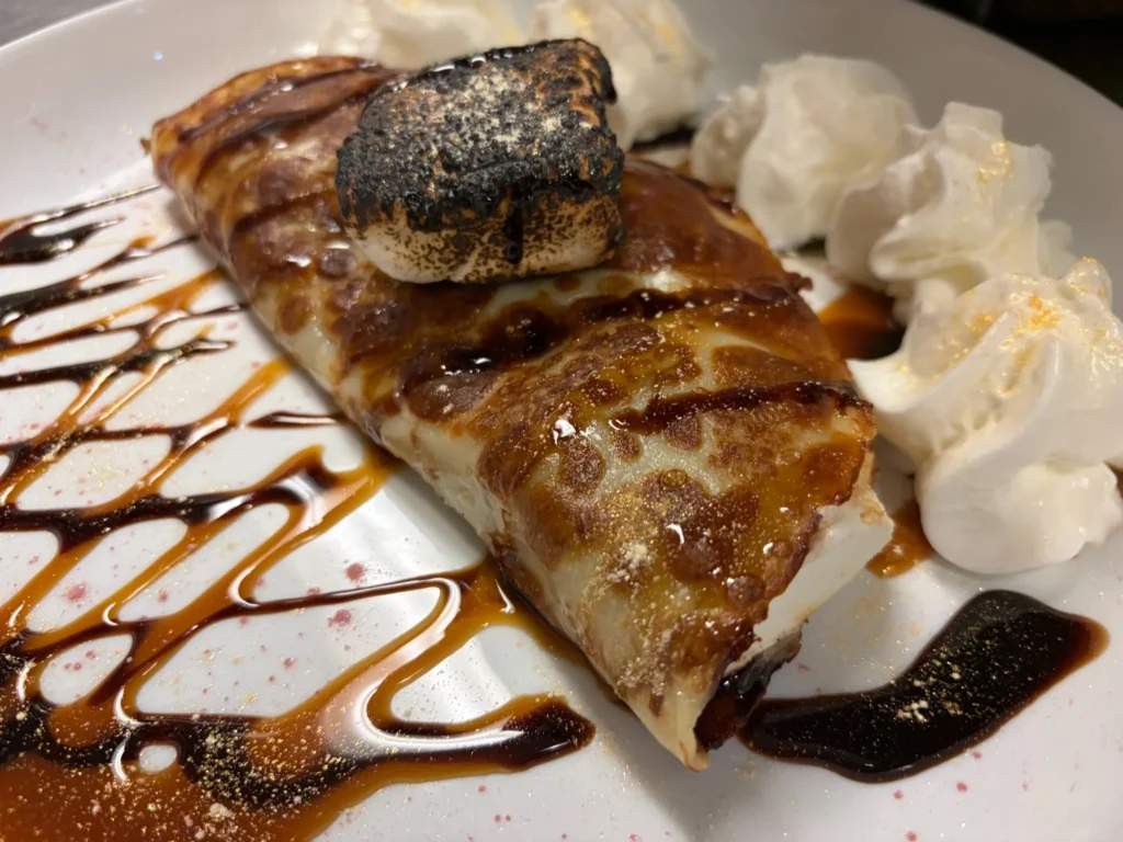 Close-up of a dessert crepe with toasted marshmallow, whipped cream, and chocolate-caramel drizzle on a white plate at Kat’s Café & Restaurant.