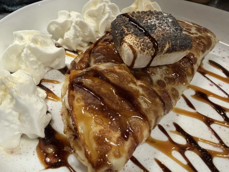 Dessert crepe filled with Nutella and topped with a toasted marshmallow and served with whipped cream, chocolate drizzle, and caramel on a white plate at Kat’s Café & Restaurant.